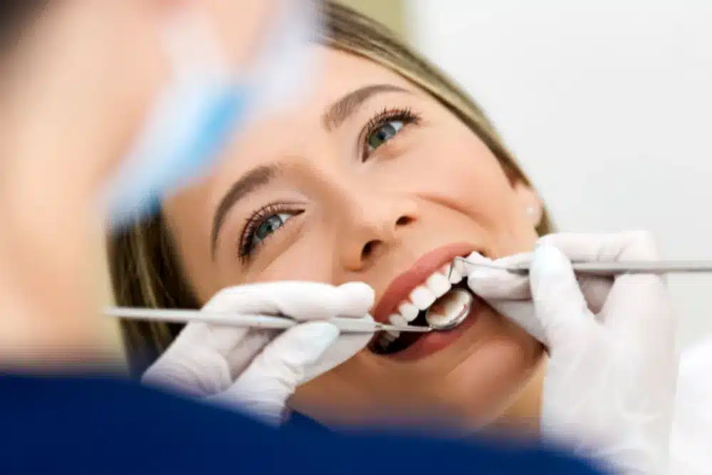 Patient smiling during a dental exam while the hygienist checks teeth with a mirror and probe.