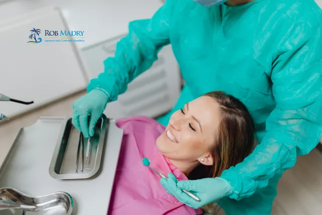 A woman laying in dentist chair with a pink dental napkin on, smiling while the dentist holds instruments near her face.