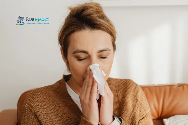 Woman blowing her nose with a tissue while sitting on a couch, highlighting the connection between oral health and immune system wellness.